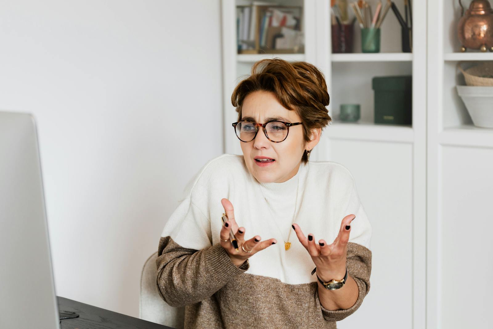 Expressive middle aged woman in casual outfit and eyeglasses gesticulating while having video conversation on computer in modern office